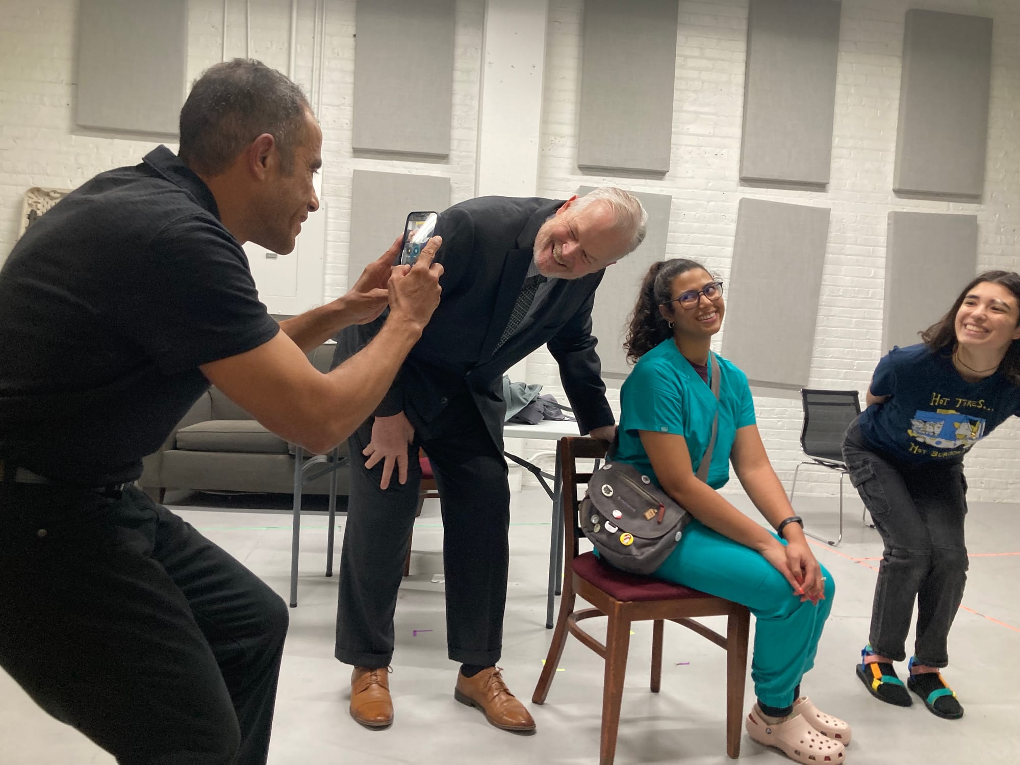 A man crouches while taking a photo with his phone of three people posing playfully in a bright room with white brick walls and acoustic panels. 